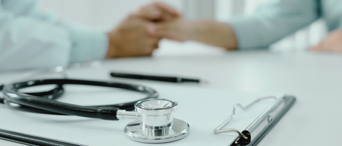 A stethoscope rests on a clipboard in the foreground as two people hold hands across a white table.