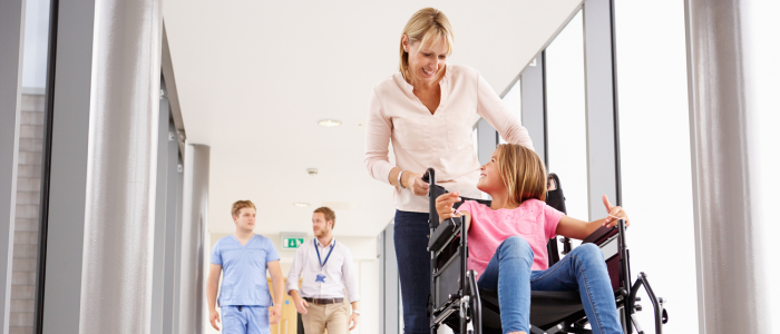 A woman pushes a child in a wheelchair down a hospital corridor while two clinicians walk behind them.