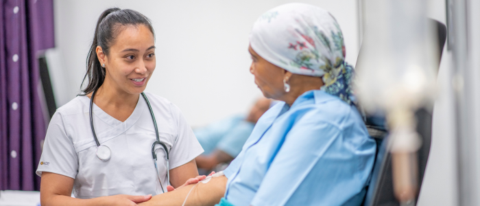 Nurse with stethoscope speaks with a patient wearing a headscarf while receiving treatment in a clinical care setting.