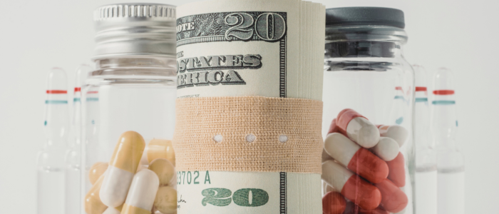 Rolled-up twenty-dollar bill wrapped with a bandage, placed between glass jars filled with capsules and ampoules, symbolizing the high cost of healthcare and medication.