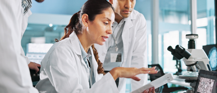 Scientists in lab coats review tablet data beside a microscope in a clinical research laboratory.