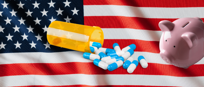 US flag with an overturned pill bottle, scattered tablets, and a pink piggy bank, illustrating prescription drug costs.