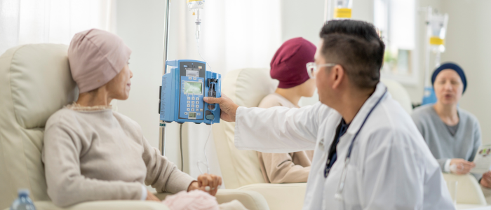 Doctor adjusts IV infusion pump beside seated patient wearing headscarf in a hospital chemotherapy treatment room.
