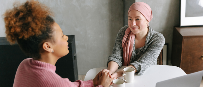 Woman holds hands with cancer patient wearing headscarf as they talk over coffee.