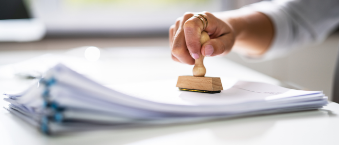 A hand holding a wooden stamp presses down on a stack of papers on a desk.