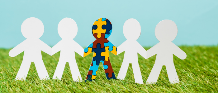 Paper human figures holding hands on grass, with the central figure covered in colourful puzzle pieces representing autism.