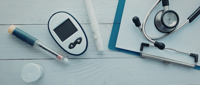 Glucose meter, insulin pen, syringe, and stethoscope arranged on a wooden surface with a medical clipboard.