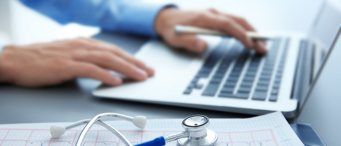 A close-up of hands typing on a laptop beside a stethoscope and medical chart on a desk.