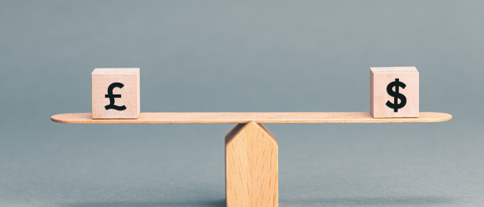 A wooden balance scale with a pound sterling (£) block on one side and a US dollar ($) block on the other, symbolising equal drug pricing in the UK and US.