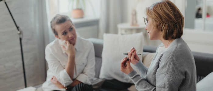 Two women sit on a couch having a focused conversation in a bright living room setting.