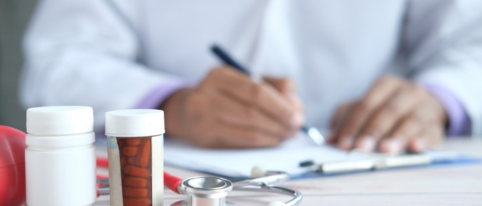 A blurred figure writing on a form with healthcare items, including pills and a stethoscope, in the foreground.