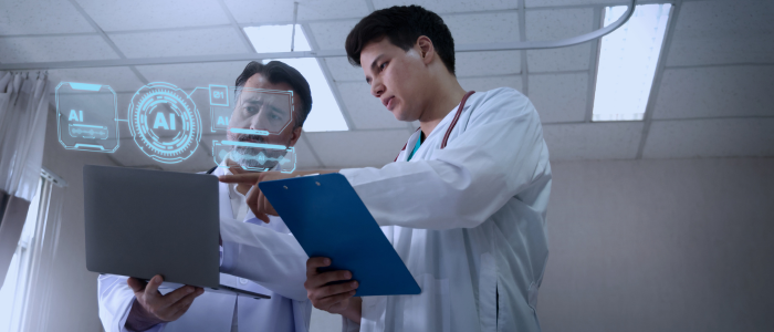 Two clinicians review a laptop and clipboard in a hospital room with holographic AI interface graphics above the screen.