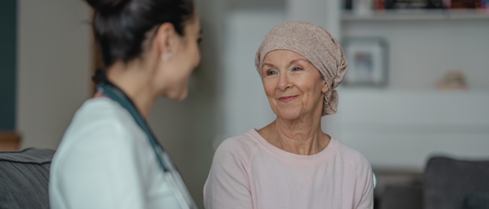An older woman wearing a headscarf smiles while speaking with a clinician seated beside her in a living room.
