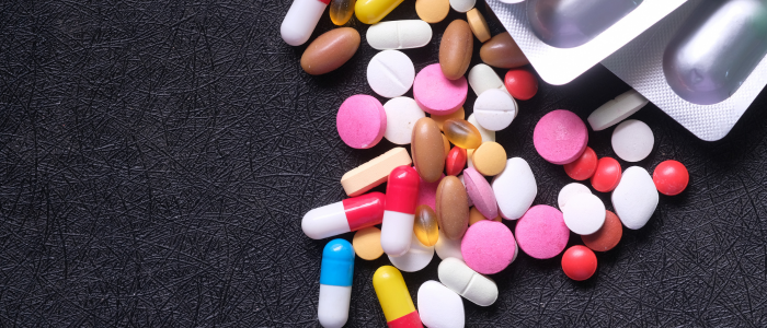 Mixed assortment of colourful pills, tablets, and capsules scattered beside blister packs on a dark surface.