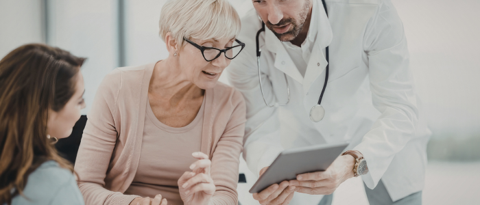 Doctor shows tablet to older woman and younger woman during consultation, discussing medical information together.