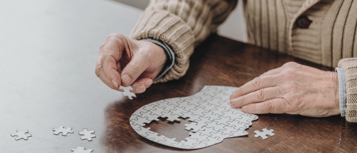Elderly hands assemble incomplete puzzle shaped like a human head, missing pieces in the area of the brain. To represent that GSK, UK DRI and HDR UK to explore link between shingles vaccine and reduced dementia risk in real-world data study.