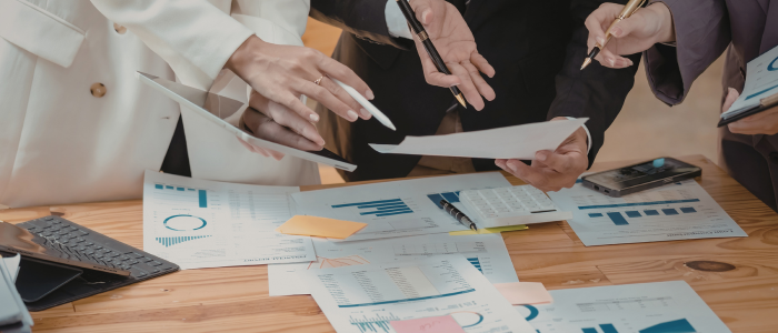 People gather around a table reviewing printed charts, graphs, and documents, with hands pointing and taking notes.