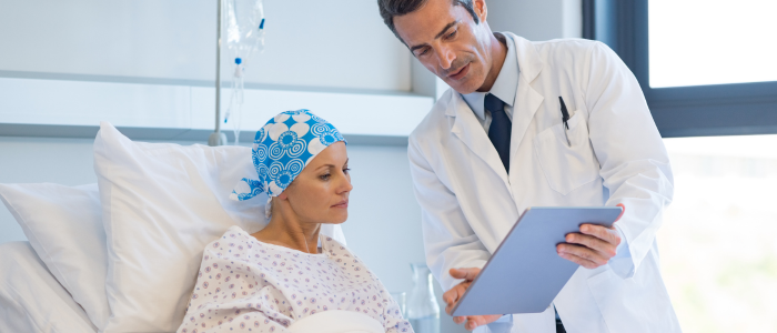 Doctor shows a tablet to a hospital patient wearing a headscarf while she sits upright in a hospital bed.