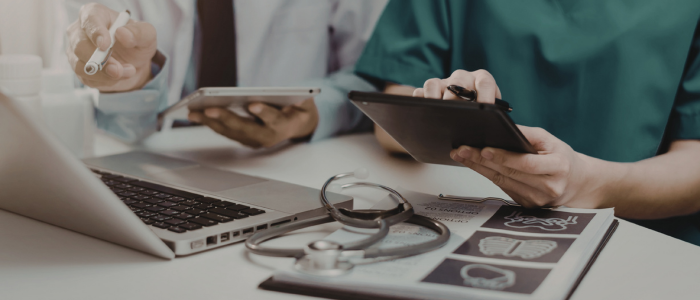 Healthcare professionals use tablets beside a laptop, stethoscope, and medical charts during a clinical discussion.