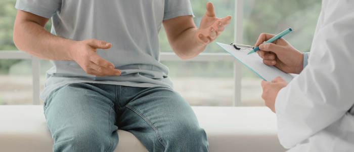 A patient sits on an examination table and gestures while a clinician holds a clipboard and writes notes during a consultation.