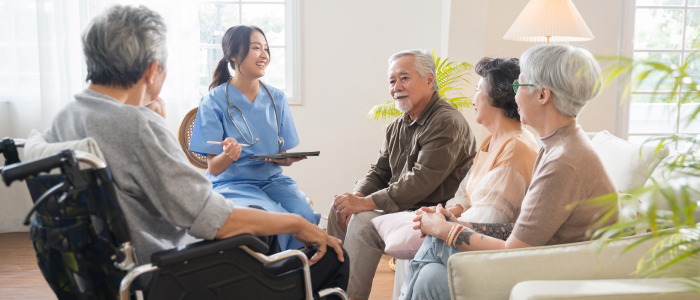 Healthcare professional in blue scrubs discusses patient experience with a seated group of older adults in a care setting.