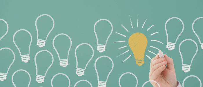 Hand drawing glowing yellow lightbulb among white outlines on green chalkboard, symbolizing innovative ideas or solutions.