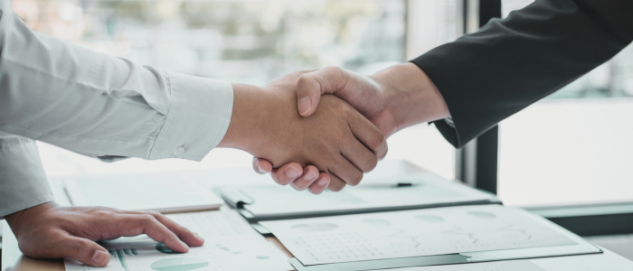 Two people shake hands over business documents on a desk.