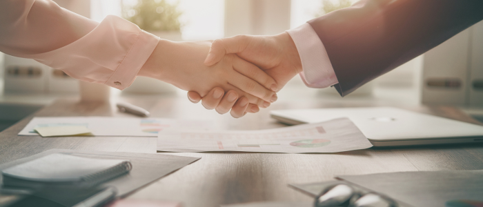 Two people shake hands over a desk with papers, laptop, and glasses in soft sunlight.