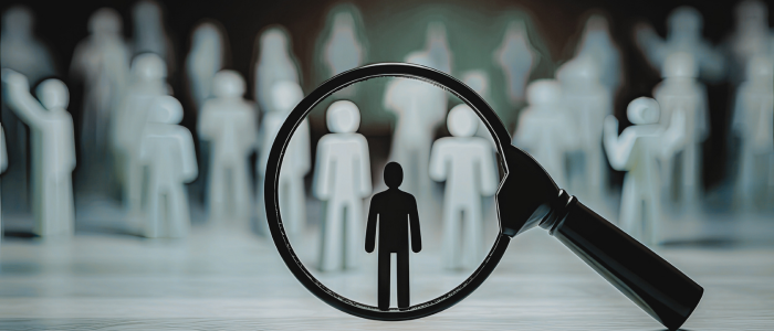 Magnifying glass highlights a single dark figure among blurred crowd of human icons on a surface.