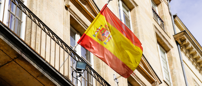 Spanish flag with coat of arms hanging from the balcony of a classic stone government building, representing national regulatory policy