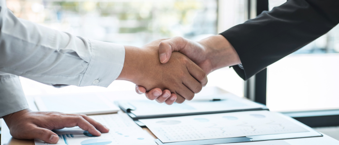 Two professionals shake hands over a desk covered with reports, charts, and documents in a business meeting.