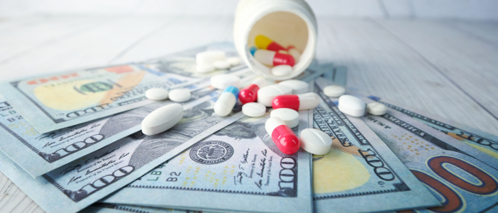 Colourful prescription pills and capsules spilling from a white bottle onto US hundred-dollar bills on a wooden surface, representing the cost of medicines under Medicare drug pricing policy.