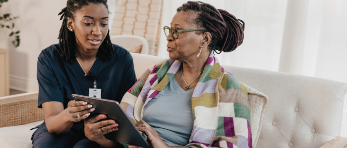 Healthcare worker shows tablet to older woman sitting on couch with colorful blanket.