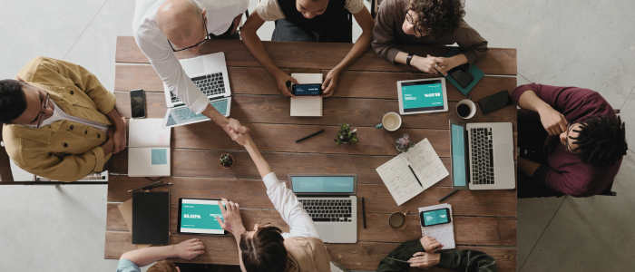 Team collaborates around table with laptops and tablets as two people shake hands.