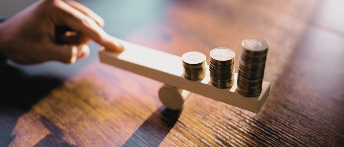 A hand balances stacked coins on a wooden seesaw, symbolizing weighing costs and value in healthcare or drug pricing decisions