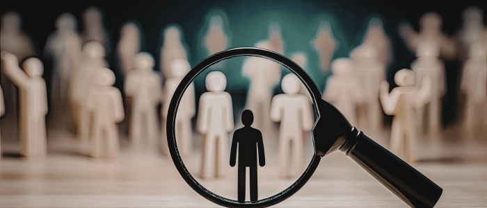 A magnifying glass highlights a single human figure among many identical figures arranged in the background.