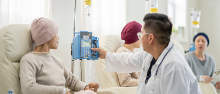 Clinician adjusts an IV infusion pump beside seated patients receiving treatment in a hospital ward.