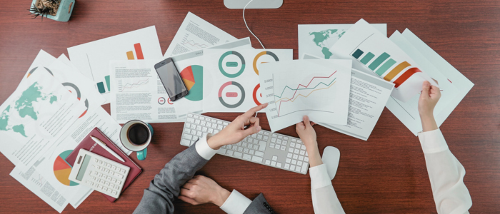 Two people review charts and graphs with data reports, maps, and coffee on a wooden desk