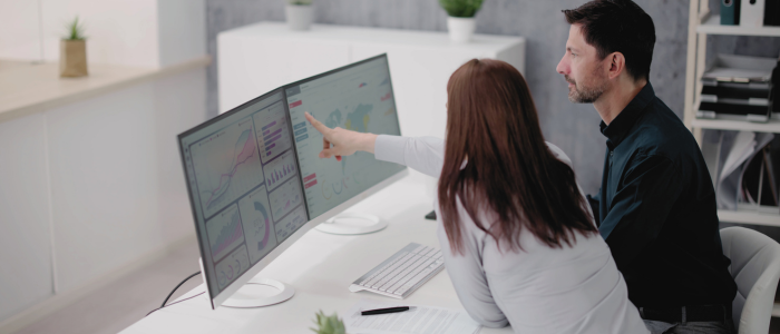 Two professionals analyzing data on dual computer monitors in an office. To represent that DATA-CAN and Arcturis form partnership to advance oncology RWE.