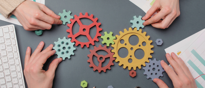 Hands assembling colorful interlocking gears on a desk beside charts and a keyboard, symbolizing collaboration and process integration.